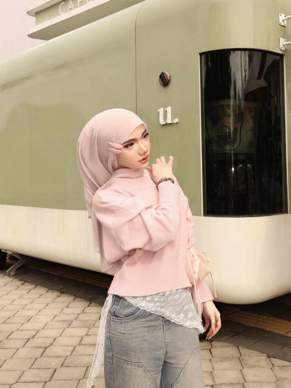 Woman in dusty pink chiffon hijab and knit sweater posing by green trailer.