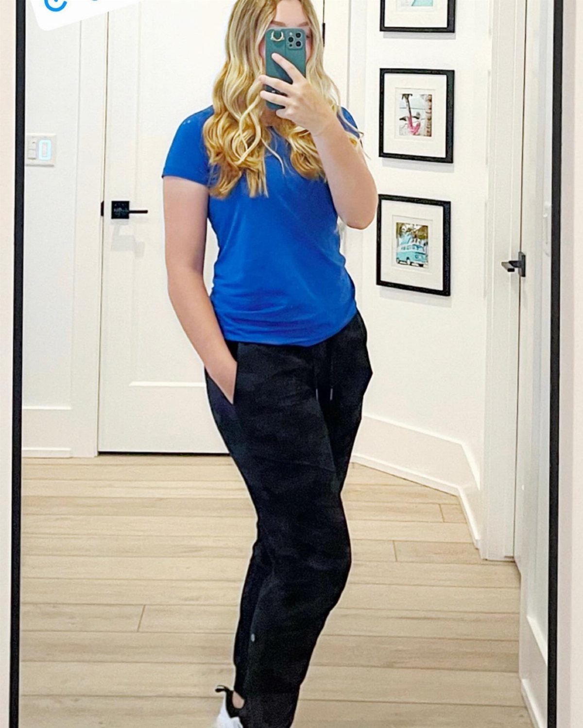 Woman in blue t-shirt and black athletic pants taking a mirror selfie indoors.